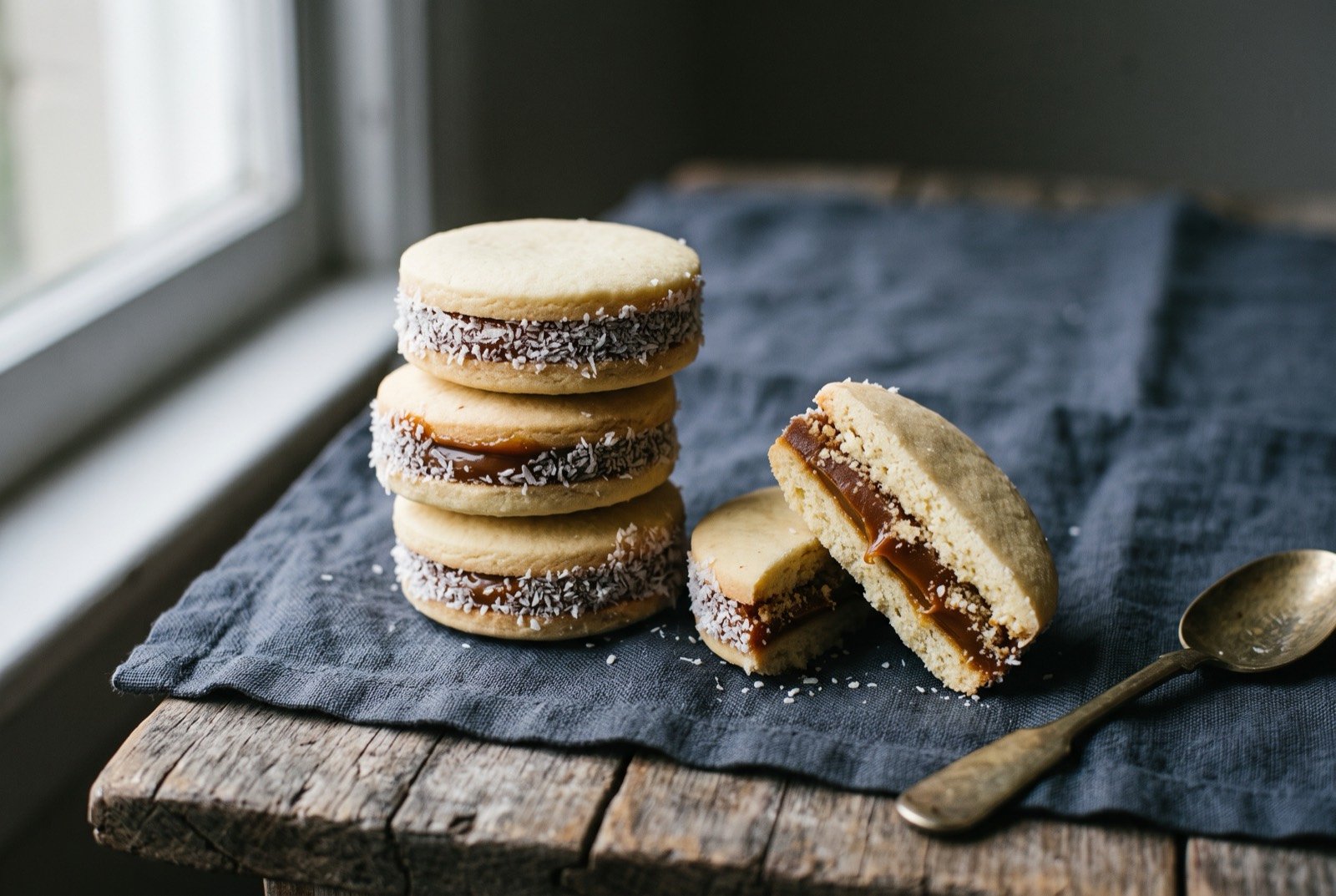 Alfajores, shortbread biscuits sandwiched with dulce de leche and rolled in coconut: a row of small round biscuits sandwiched with a thick, glossy layer of dark caramel coloured dulce de leche oozing slightly at the edge.