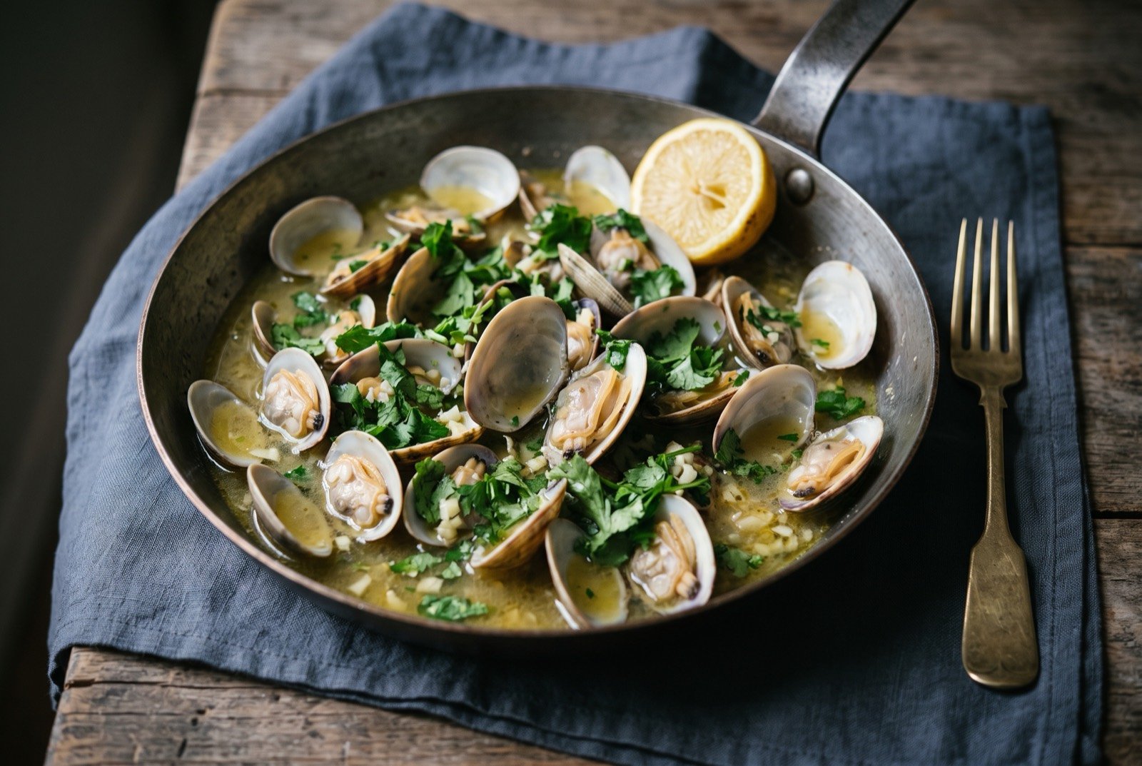 Amêijoas à Bulhão Pato, clams in garlic and coriander: a wide shallow pan of open clam shells sitting in a thin pale golden garlic and olive oil broth, torn green coriander scattered generously across.