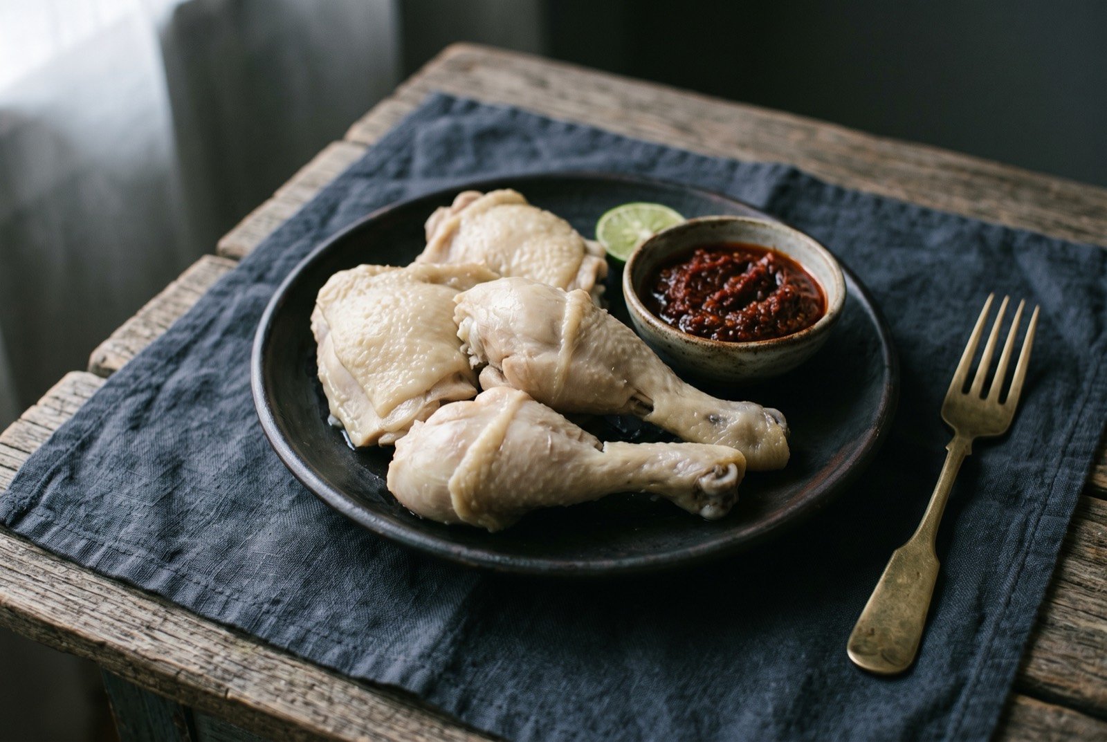 Ayam pop, pale poached and briefly fried chicken: pale, un-browned pieces of jointed chicken on a plate, notably lighter than typical fried chicken, a small dish of dark red sambal resting alongside for contrast.