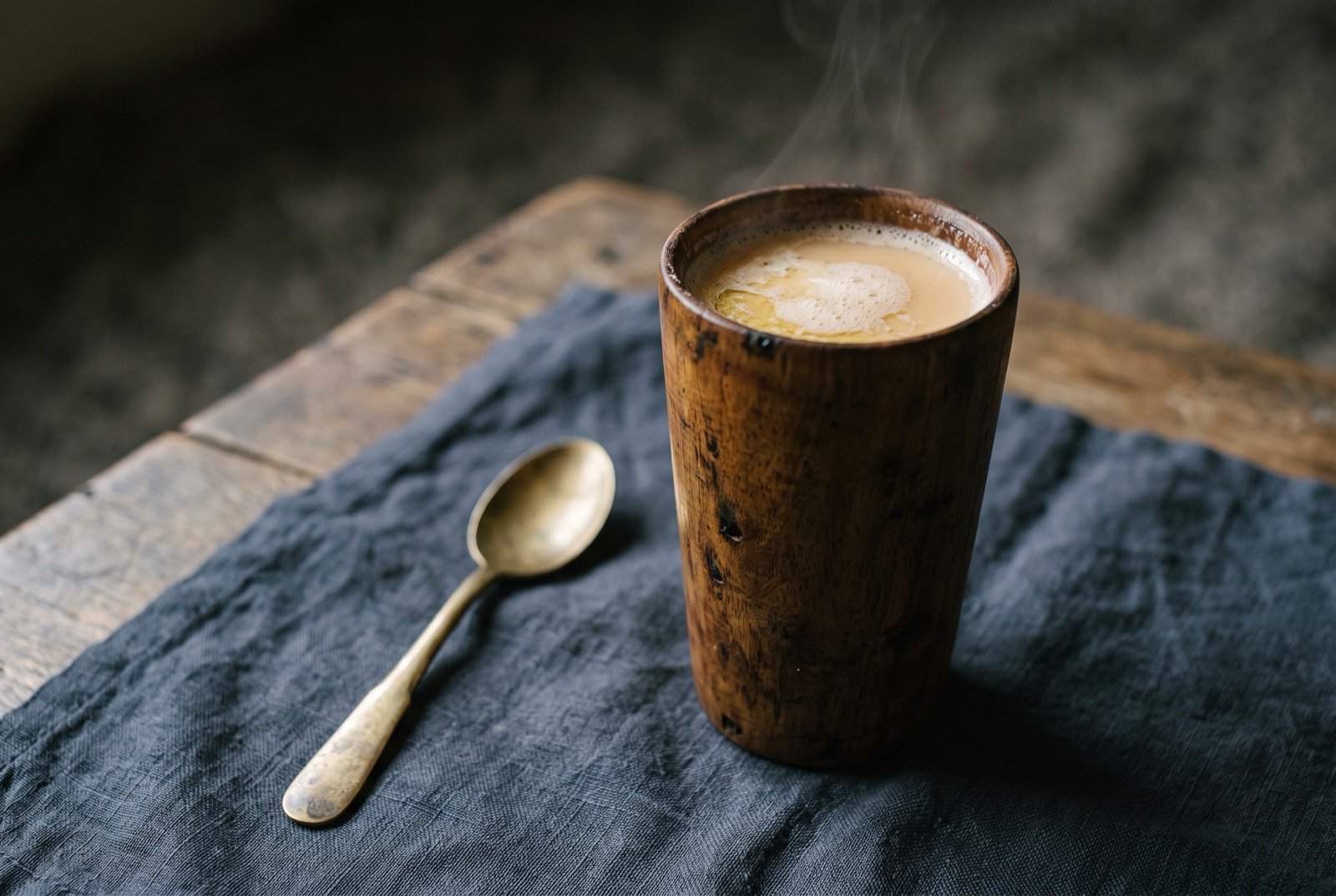 Butter tea, suja and gur gur cha, churned salted tea and yak butter: a tall, narrow wooden cup filled with pale, faintly frothy amber tea, a thin oily sheen visible at the rim where the churned butter has emulsified.
