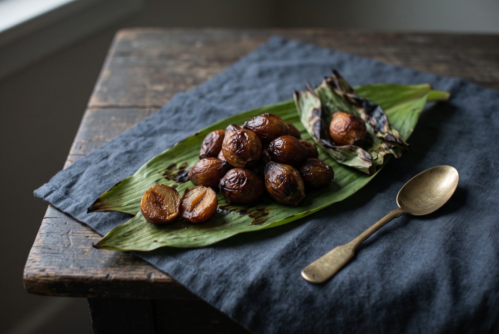 Camas bulb pit-roast, small sweet bulbs slow-roasted underground for a day: a small heap of dark, caramel brown roasted bulbs, glossy and slightly collapsed, resting on a large wilted green leaf, a few unwrapped from their charred leaf parcels beside them.