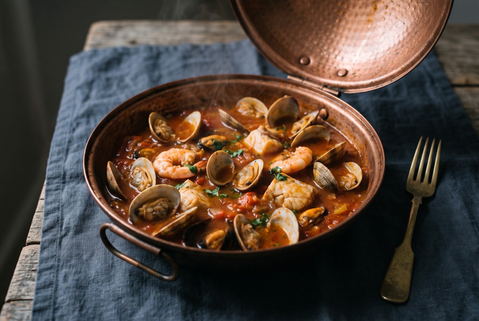 Cataplana de marisco, sealed shellfish and fish stew: an open hinged copper cataplana pan seen from above, clams still in their shells, pink prawns and pale flakes of fish sitting in a rust red tomato and pepper broth.