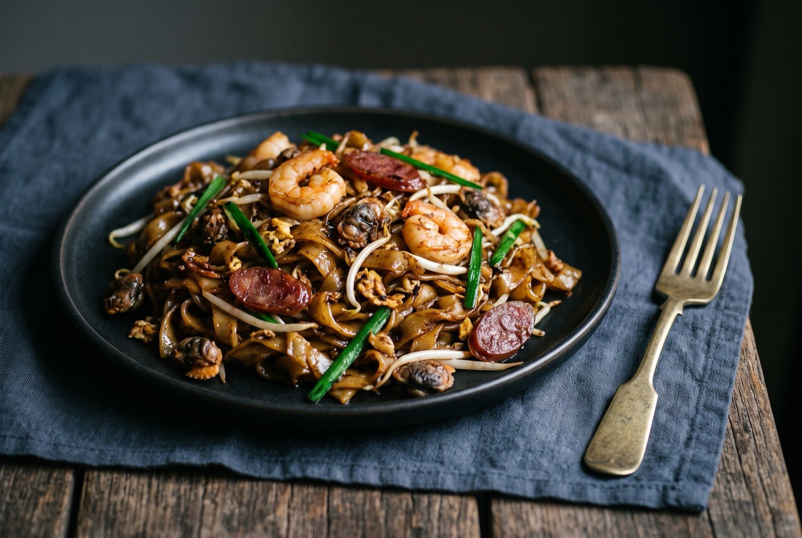 Char kway teow, flat rice noodles wok-fried with egg, prawns, cockles and Chinese sausage: a dark plate of glossy, charred flat rice noodles studded with pink prawns, dark cockles, slices of red Chinese sausage and scrambled egg.