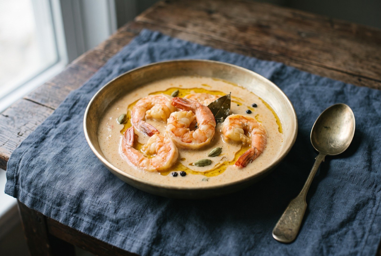 Chingri malai curry, prawn in a coconut milk sauce: large pink-orange prawns half submerged in a pale, creamy coconut coloured sauce, a few whole green cardamom pods and a bay leaf visible at the surface.