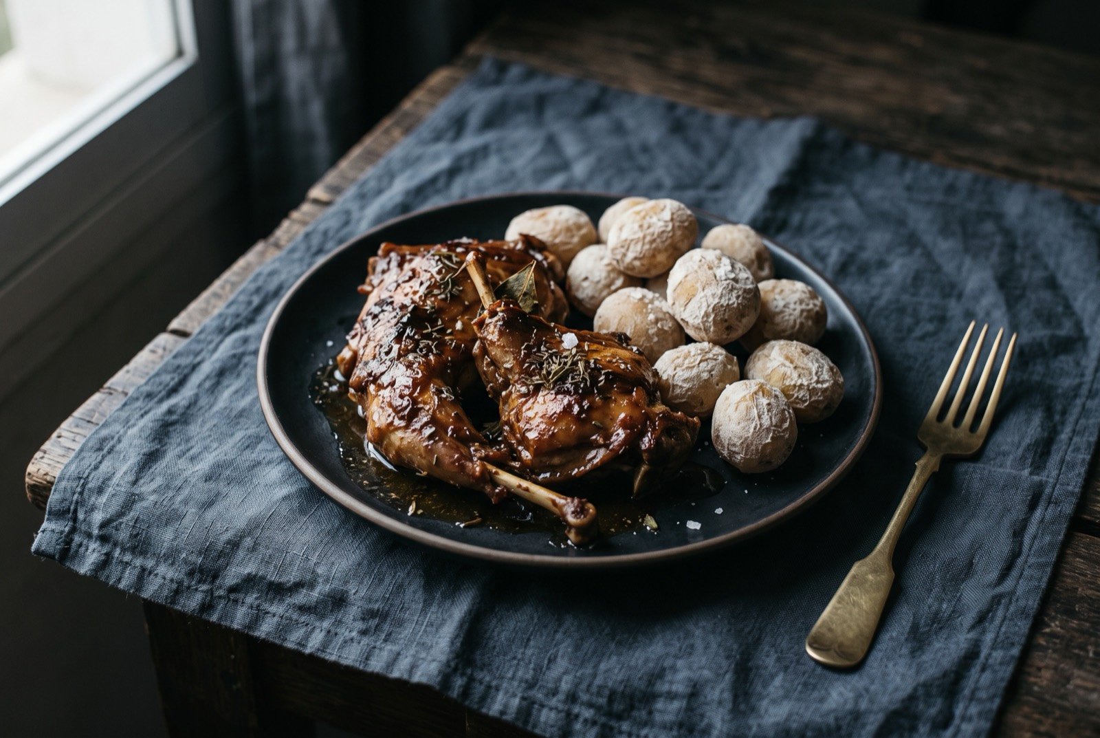 Conejo al salmorejo canario, wine-marinated rabbit: deeply browned, bone-in rabbit pieces glazed with a dark reduced marinade, arranged on a dark plate beside a small pile of wrinkled salt-crusted potatoes.