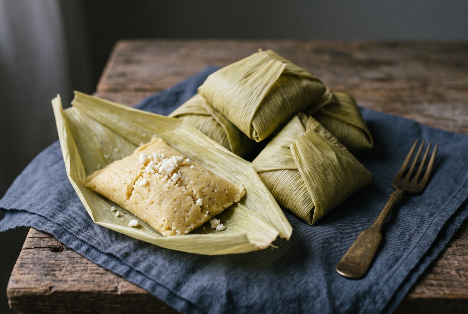Corundas, a triangular corn tamale wrapped in maize leaves: a cluster of pale green maize leaves, folded into distinct angular, many sided parcels, one leaf peeled back to reveal a dense.