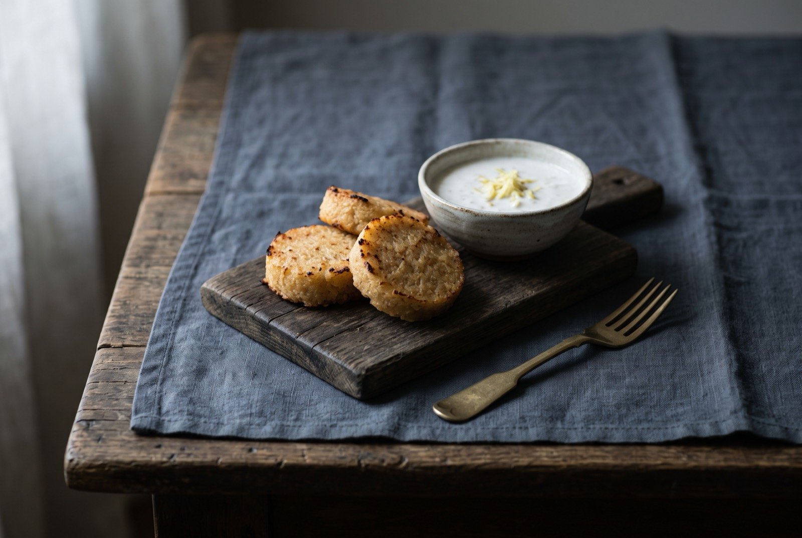 Delul diokang, Palau's smoked grated tapioca cakes with coconut dip: small firm golden-brown tapioca cakes with lightly charred edges, arranged in a loose row on a dark board beside a small bowl of white coconut dip flecked with grated ginger.