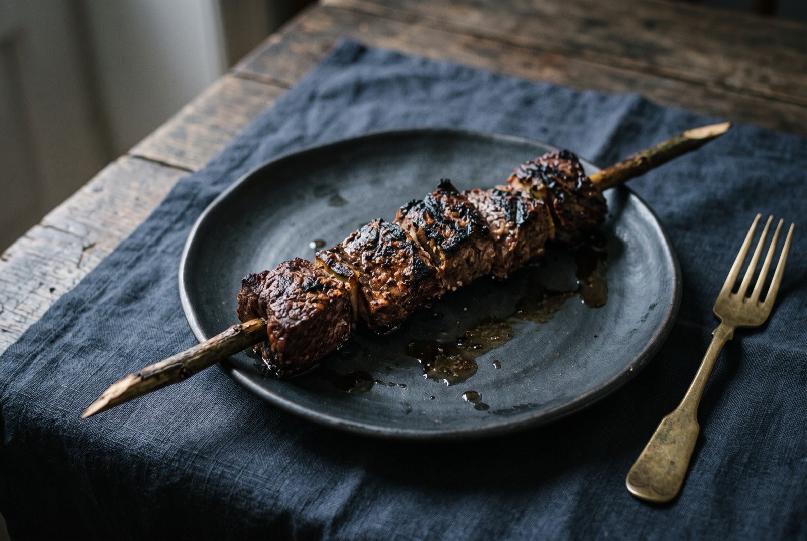 Espetada em pau de louro, beef skewered on fresh bay laurel: large charred cubes of beef threaded on a stripped fresh bay laurel branch, deeply seared outside, resting across a dark plate with visible drips.