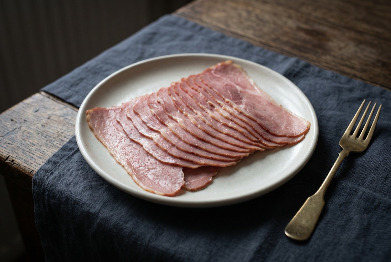 Hangikjöt, smoked lamb sliced thin for Christmas: thin pale pink slices of smoked lamb fanned across a plain plate, a faint smoky sheen catching the light, nothing else in the frame.