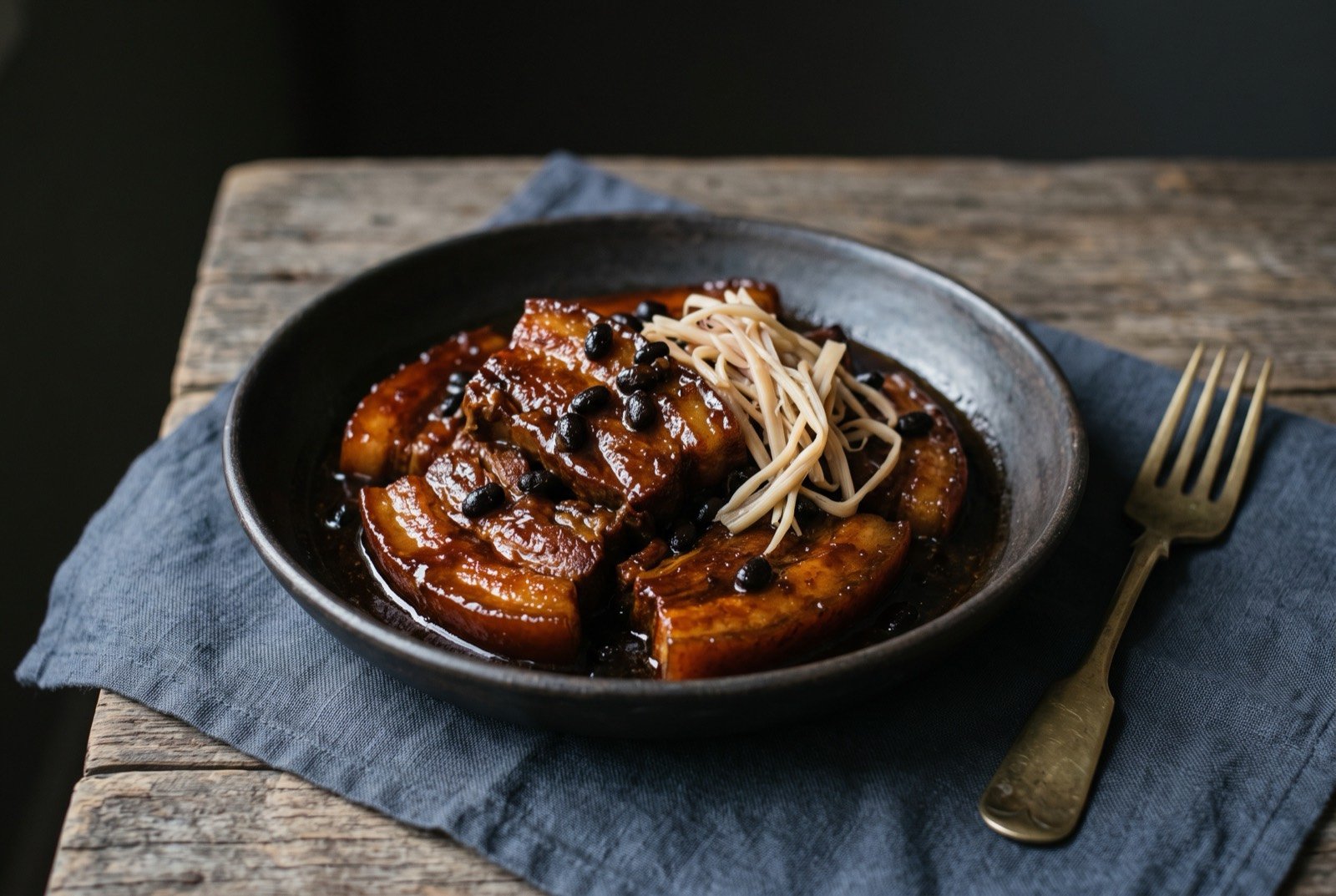Humba, pork belly braised sweet and savoury with fermented black beans and banana blossom: a dark shallow bowl of glossy, deeply caramelised pork belly slices in a thick dark brown reduced sauce, scattered with whole fermented black beans.