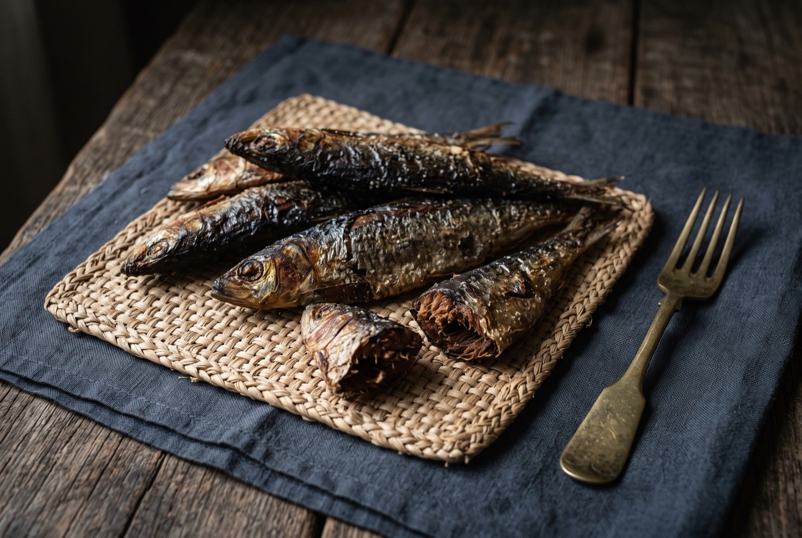 Kethiakh, braised and sun dried sardinella: whole small dried fish, dark brown and leathery, skin blistered and salt crusted, laid out on a woven raffia mat, a few pieces broken open to show dense dark flesh.