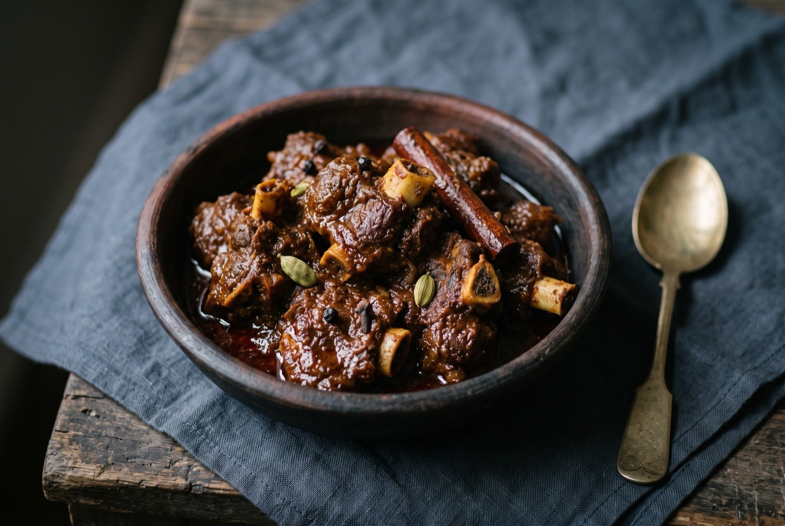 Kosha mangsho, mutton reduced dark and dry with whole spice: bone-in mutton pieces coated in a thick, deep reddish-brown, glossy sauce that clings tightly rather than pooling, a few whole cloves.