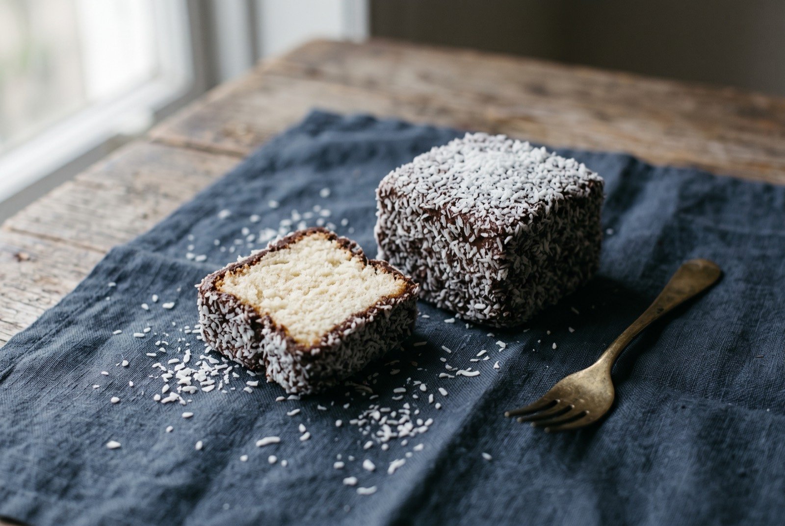 The lamington, sponge cake dipped in chocolate icing and rolled in coconut: a single square of sponge cake coated in a thin, matte dark chocolate icing and rolled evenly in white desiccated coconut.
