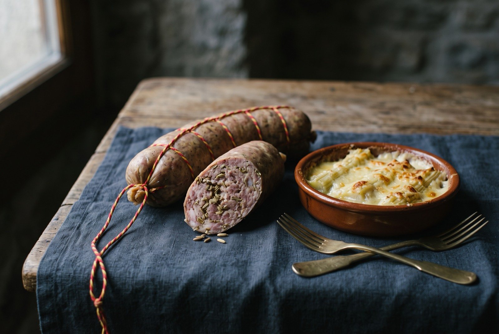 Longeole, with cardon gratin, a coarse fennel-seasoned Genevan pork sausage with a gratin of spined thistle: a thick pale sausage cut into rounds on a plate, one segment split open to show its coarse, fennel-flecked pink and white interior.