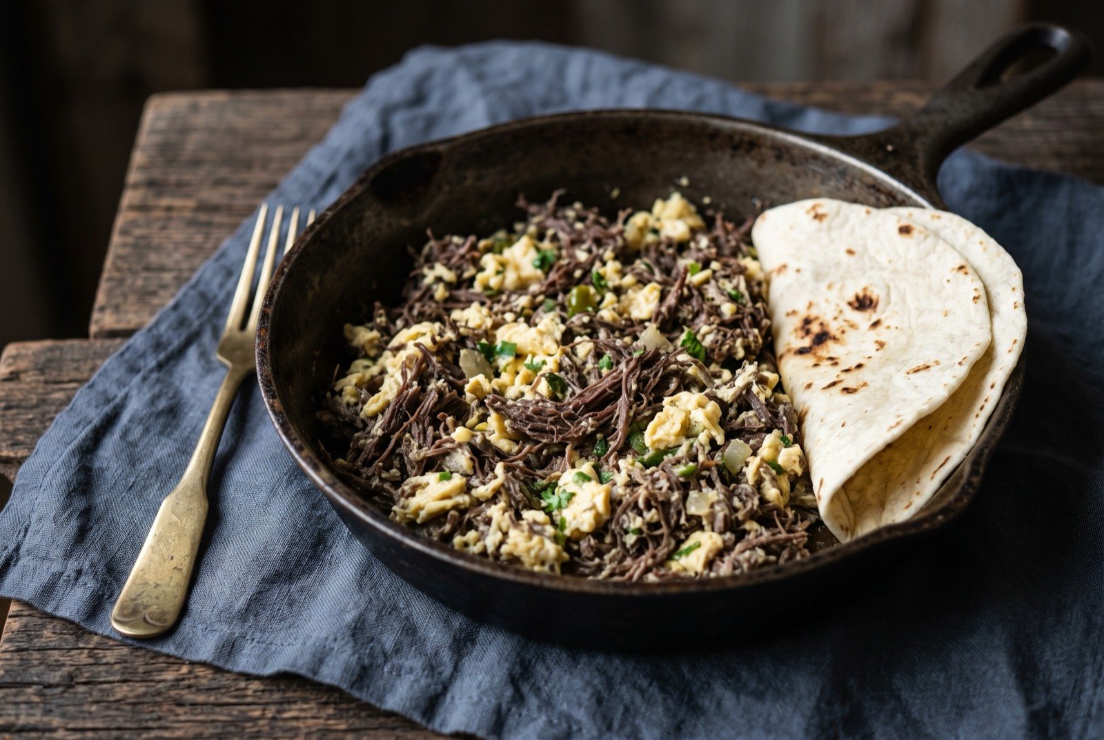 Machaca con huevo, shredded dried beef scrambled with egg: a mound of fine shredded dark beef threads scrambled through soft pale yellow egg in a well-worn pan, flecks of green chile and onion scattered through.