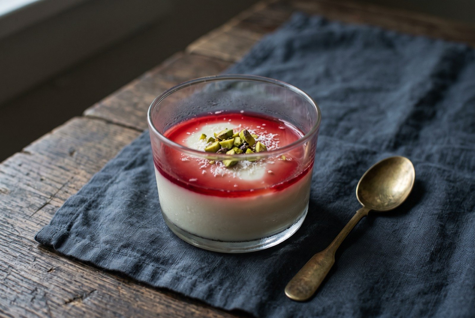 Malabi, Israel's rosewater milk pudding: a pale, softly set white pudding in a low glass, topped with a ruby red rosewater syrup pooling at the edges, a scatter of chopped pistachio and a little desiccated coconut over the top.