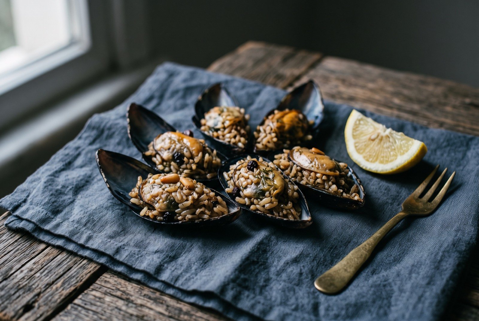 Midye dolma, mussels stuffed with spiced rice and served cold: a scattering of open mussel shells each packed with glossy, currant-flecked rice, arranged in a loose fan, a few pine nuts and flecks of dill visible in the filling.