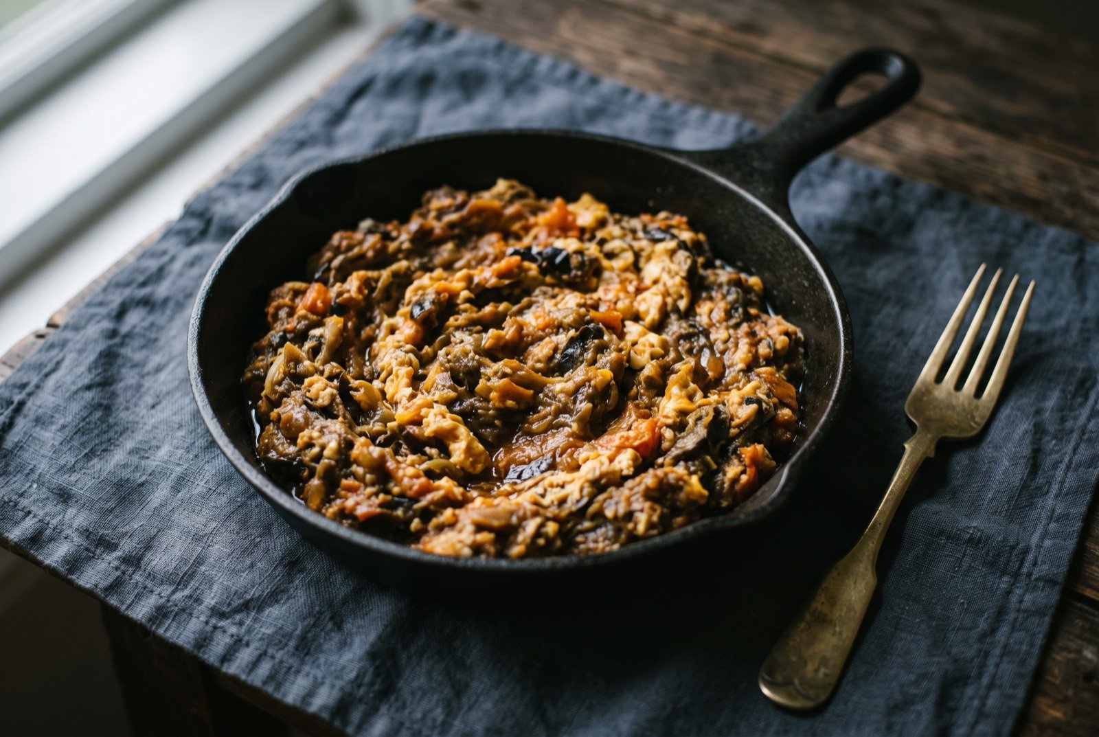 Mirza ghasemi, smoked aubergine mashed with garlic, tomato and egg into a warm savoury dip: a shallow pan of soft, deep brown mashed aubergine flecked with pale golden garlic and threads of soft scrambled egg, a thin sheen of oil pooling at the pan's edge.