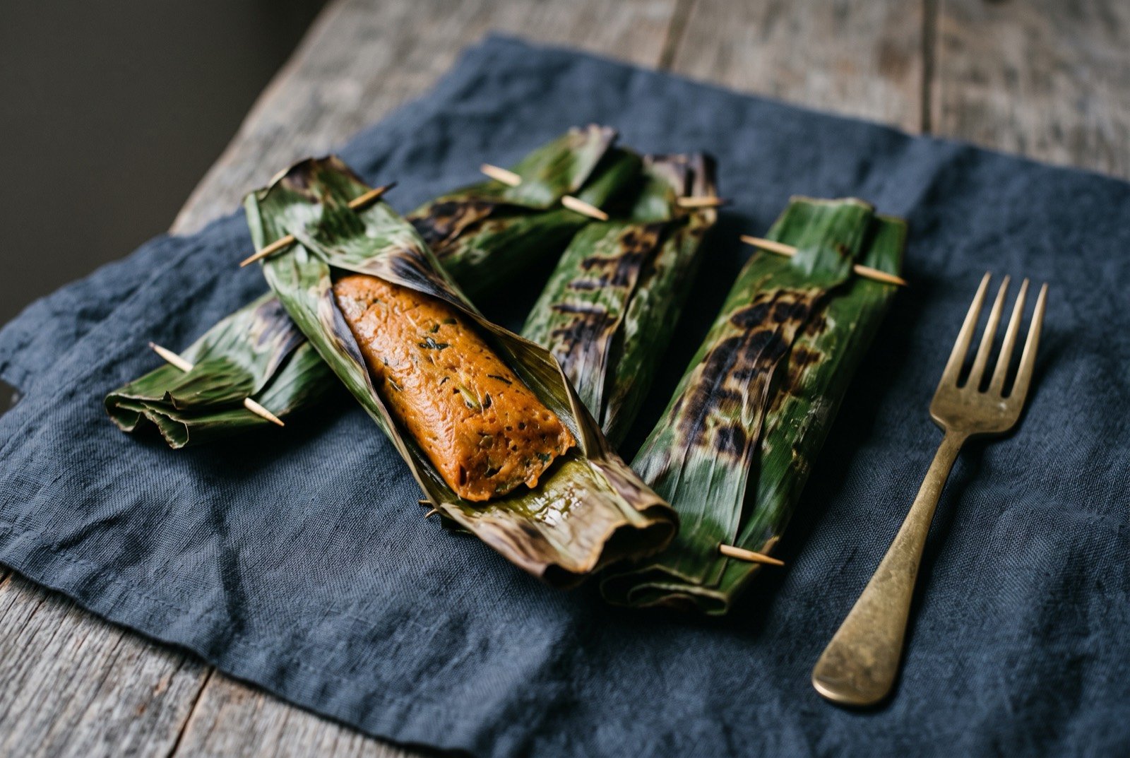 Otak-otak, spiced fish paste grilled inside a charred banana leaf parcel: a few banana leaf parcels, pinned shut with wooden toothpicks and lightly charred along their folds, one parcel unwrapped at one end to reveal a firm.