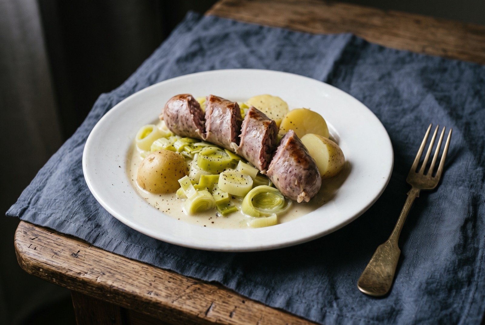 Papet vaudois, leeks and potatoes simmered with cream alongside a poached Vaudois sausage: a plate of pale, softened leeks and potatoes in loose, distinct pieces bound lightly in a creamy sauce, a plump poached grey-pink Vaudois sausage laid alongside.