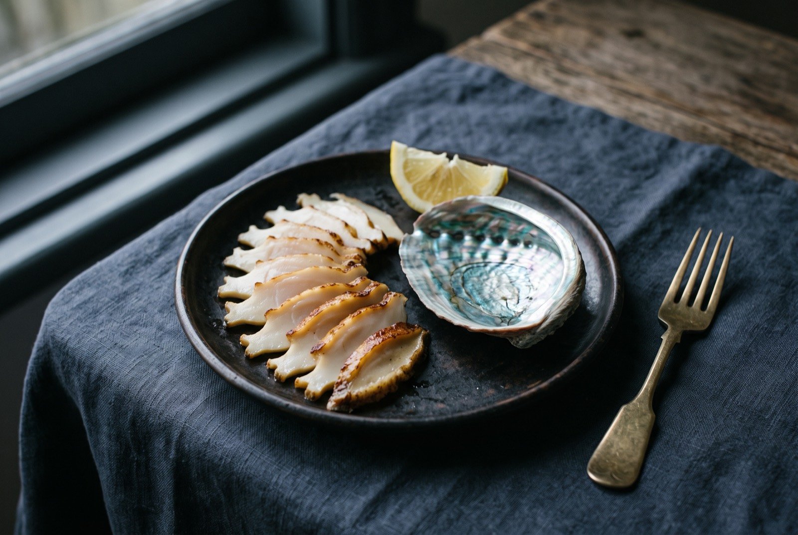 Pāua, tenderised and pan seared: thin pale ivory slices of shellfish fanned across a dark plate, each slice showing a faint seared edge, a single iridescent blue green pāua shell resting empty beside them.