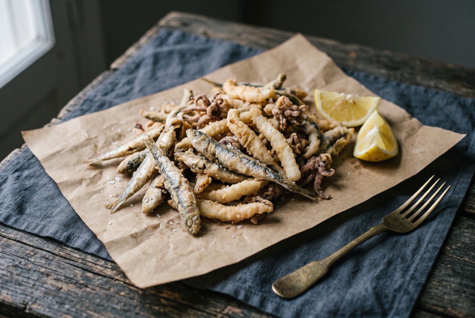 Pescaíto frito, mixed fried small fish, Cádiz style: a rustic pile of small whole fried fish and cuttlefish strips, pale golden and lightly crisped, dry rather than greasy, heaped loosely on a sheet of kraft paper.