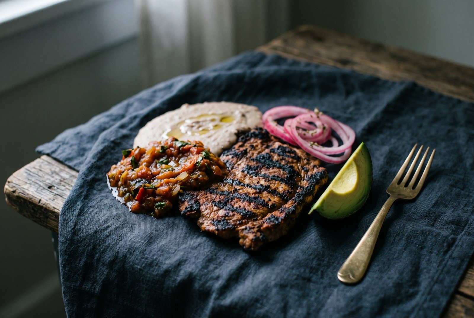 Poc chuc, sour orange marinated pork grilled over charcoal: a thin, deeply charred grilled pork steak with dark grill marks, laid beside a spoonful of charred tomato and onion chiltomate salsa and pale strained black bean purée.