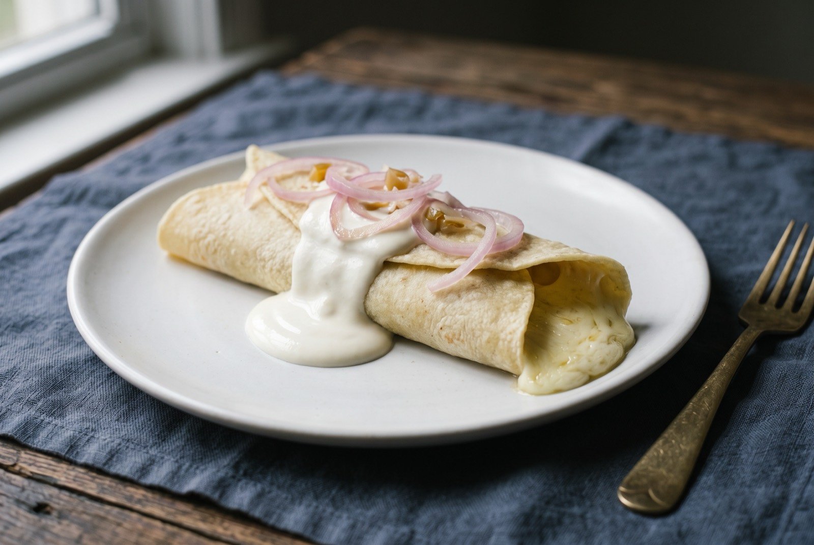 Quesillo nicaragüense, a warm tortilla wrapped around melted cheese with cream and pickled onion: a pale, softly rolled corn tortilla parcel with melted white cheese just visible at one open end, a generous pour of thick cream running down its side.