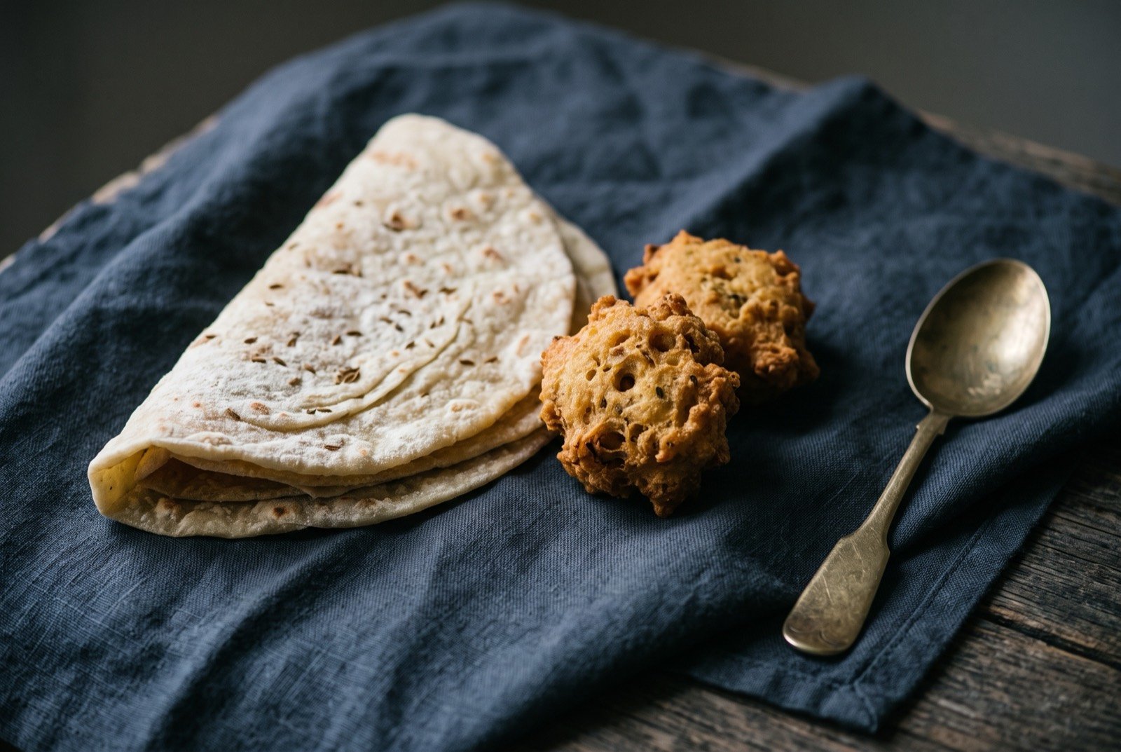 Roti and bara, laminated flatbread with split pea fritters: a folded, faintly layered flatbread dusted with flour, resting beside two or three small golden fried bara fritters with a craggy surface.