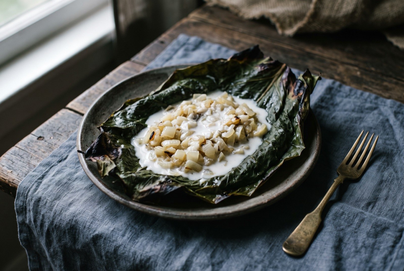 Rukau, Cook Islands cooked taro leaf with onion and coconut cream: a wrapped parcel of dark green taro leaf opened flat on a plate, filled with soft diced onion and pooled with thick white coconut cream.