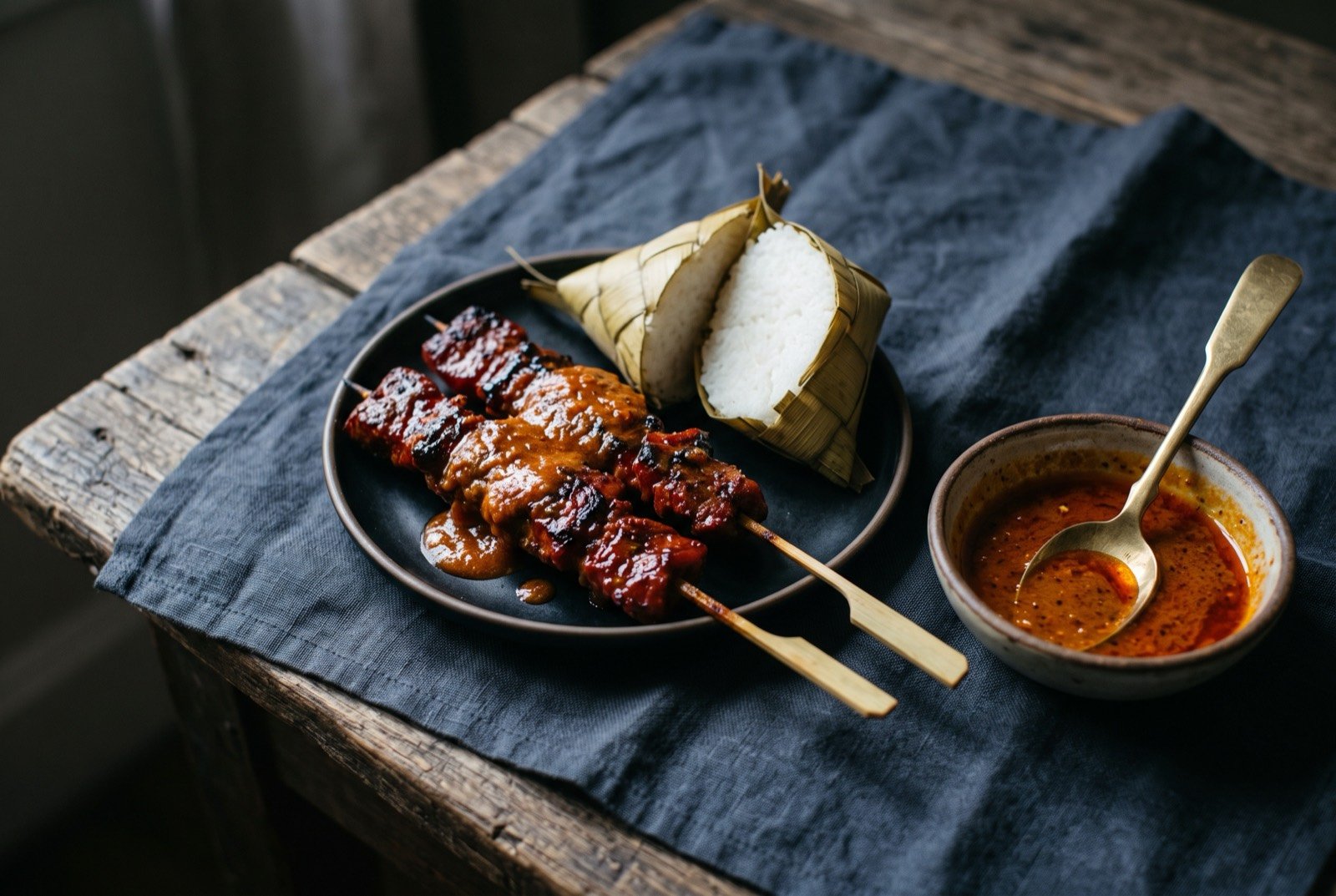 Satti, Tausug grilled skewers with puso rice and a sweet-spicy sauce: two skewers of charred, deep-red-glazed grilled beef resting across a small plate beside a diamond-shaped woven coconut-leaf puso rice pouch cut open to show plain white rice within.