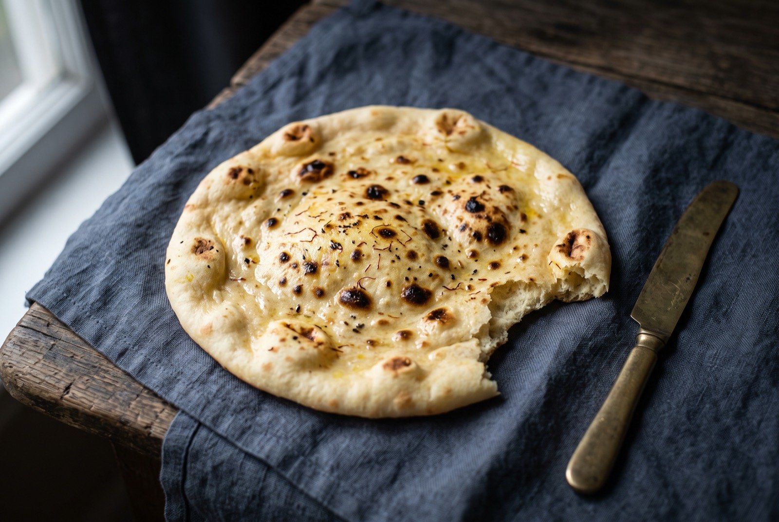 Sheermal, Lucknow's saffron and milk enriched flatbread: a round, pale golden flatbread with a faint saffron blush and dark leopard spotting across its surface, torn slightly at one edge to show a soft.