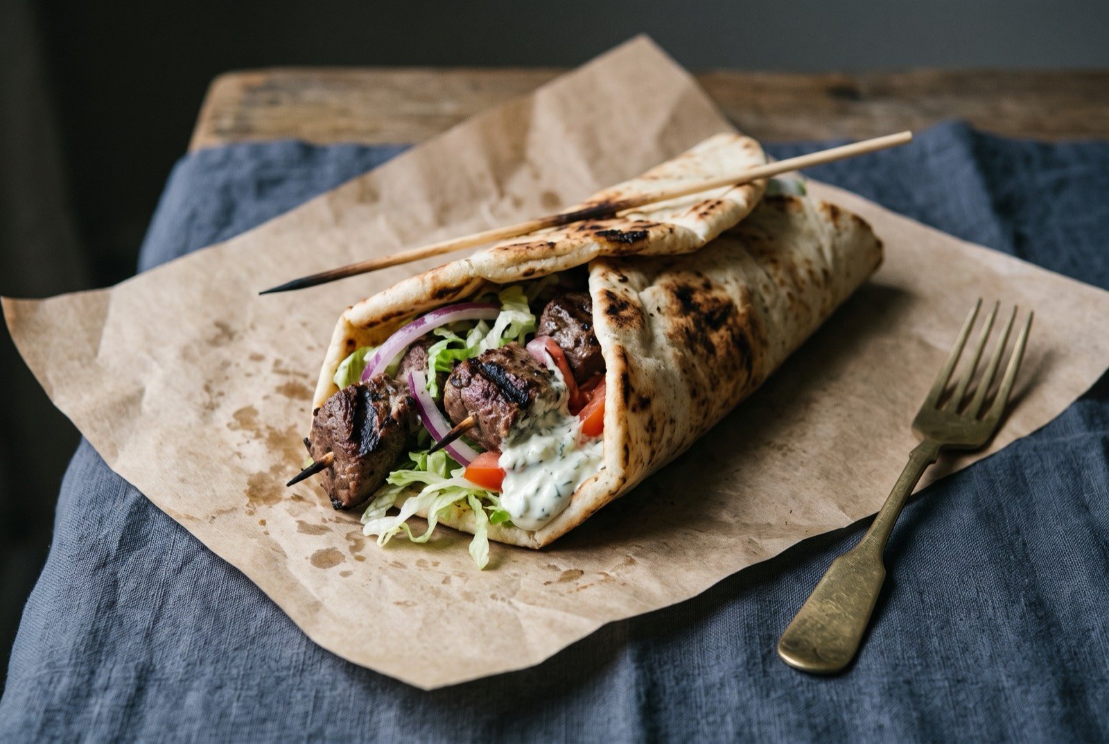 Melbourne souvlaki, skewered grilled lamb wrapped in pita with salad: a folded pita wrap resting on butcher's paper, torn open at one end to show charred cubes of grilled lamb, shredded lettuce.