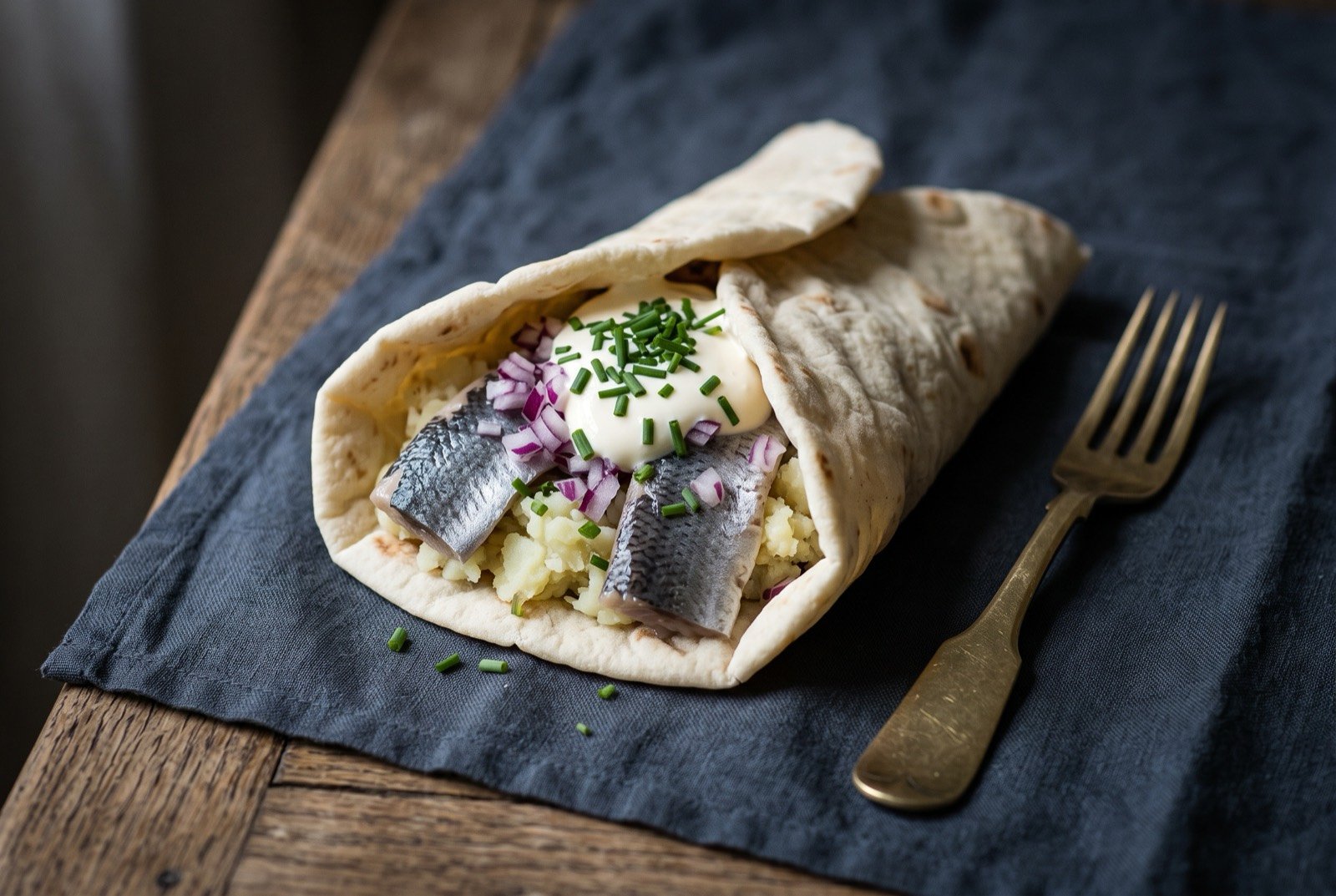 Surströmmingsklämma, fermented herring wrapped in flatbread with almond potato: a soft pale flatbread folded loosely open, revealing a filling of rough mashed potato, dark silvery-skinned fish fillets.
