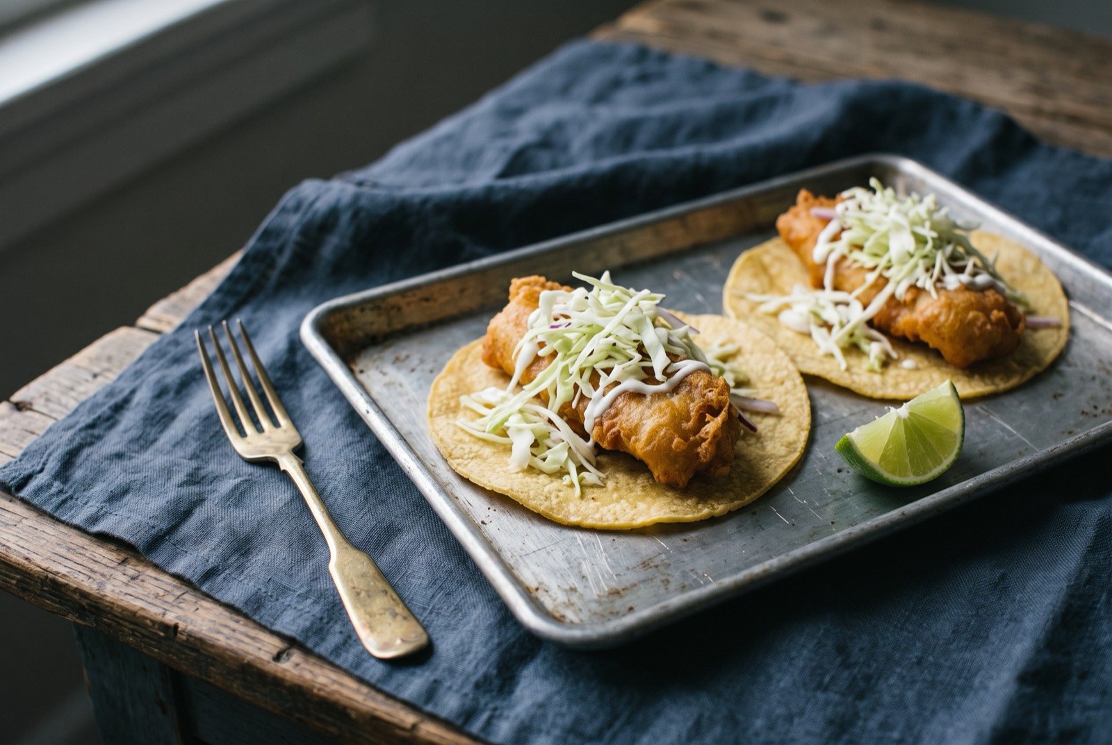 Tacos de pescado estilo Ensenada, battered fried fish tacos: two small corn tortillas each holding a piece of golden battered fried fish with a craggy crisp coating, topped with a loose pile of pale shredded cabbage.