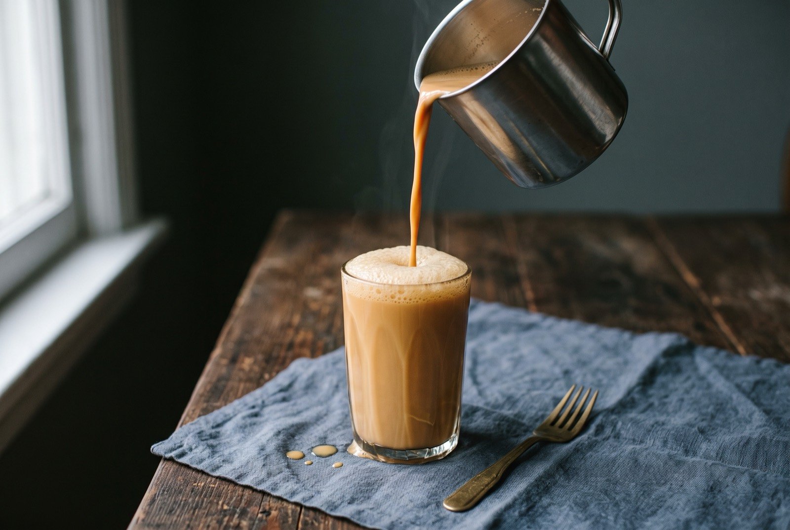 Teh tarik, pulled tea with condensed milk, poured mid-stream between two glasses: a tall glass of pale caramel-brown tea topped with a fine, stable foam, a second empty stainless steel cup resting beside it.