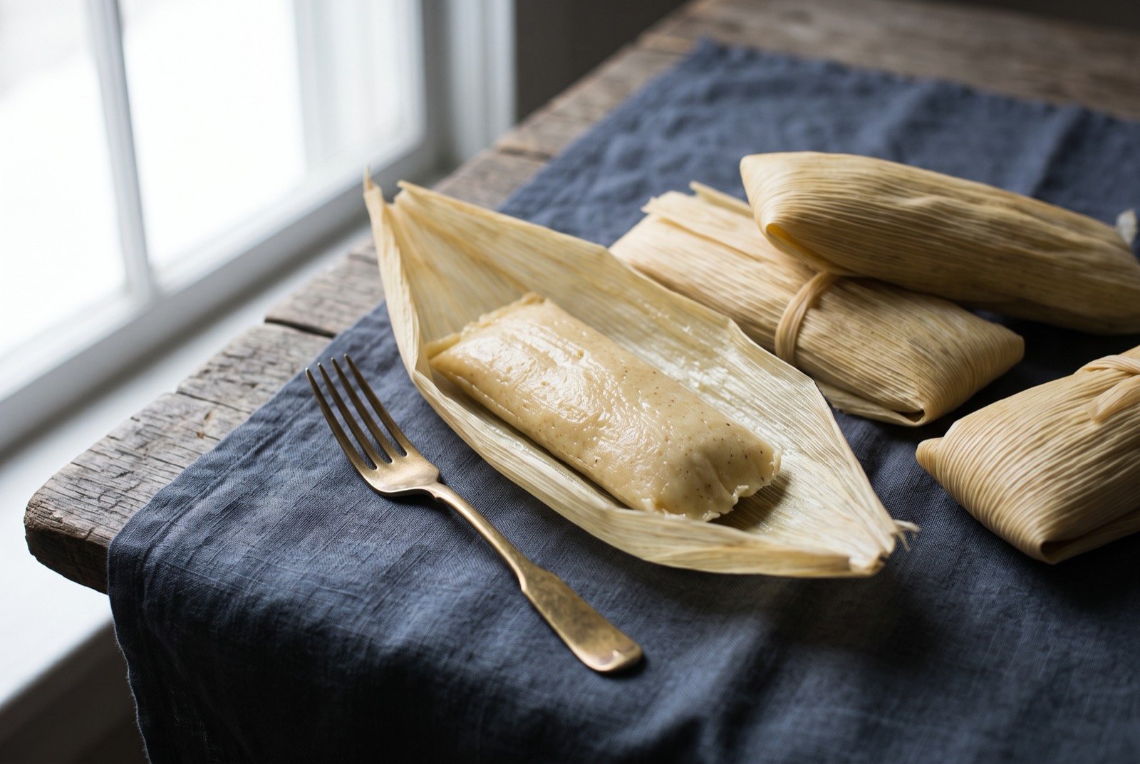 Uchepos, a soft, sweet fresh corn tamale in its own husk: pale golden fresh corn tamales still wrapped in their own pale straw coloured husks, one unwrapped to show a soft, faintly speckled.