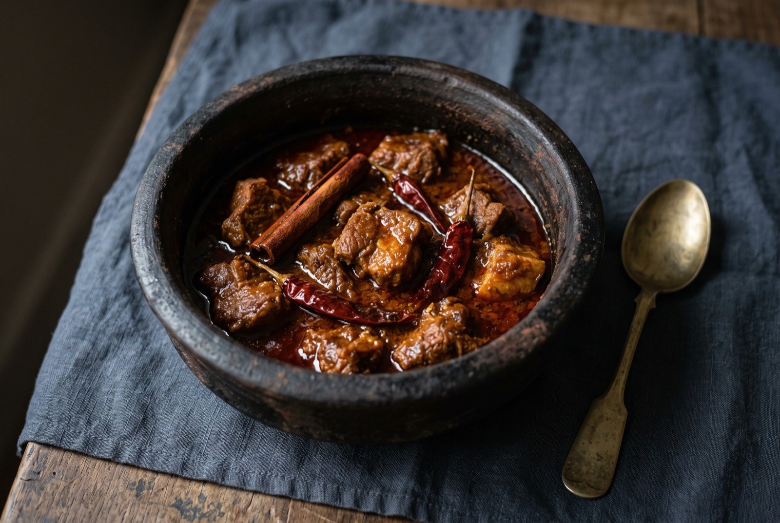 Vindalho, pork slow cooked in a dark vinegar and chilli sauce: chunks of pork in a thick, dark reddish-brown, glossy sauce inside a blackened clay pot, a cinnamon stick and a few soaked dried red chillies resting among the pieces.