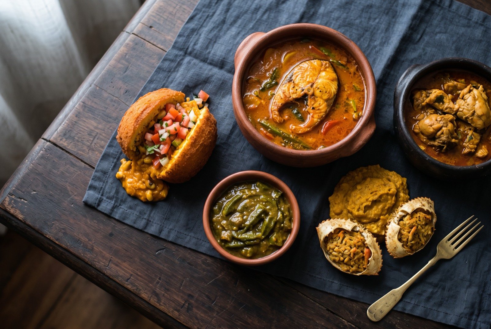 The Bahia and the Dendê Coast table: a wide dark wooden table seen from above, a split golden acarajé spilling vatapá and tomato salad beside a clay pot of burnt orange moqueca with a fish steak breaking the surface.