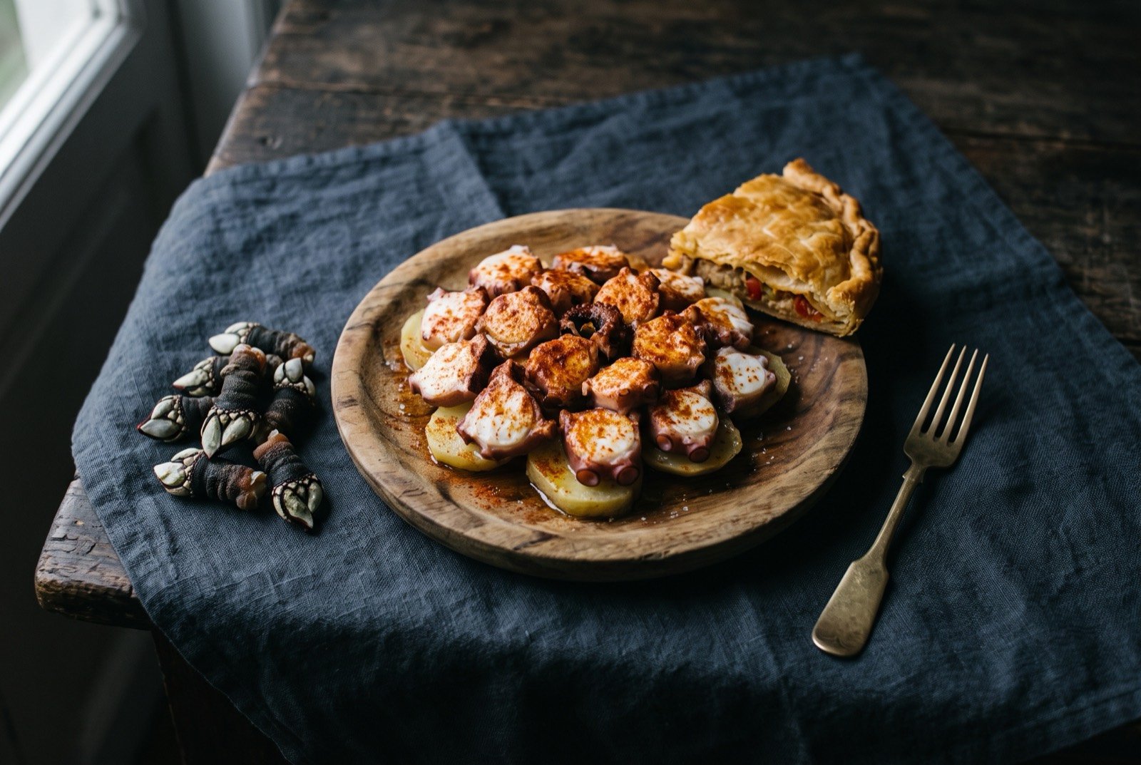 The Galicia table: a wide dark wooden table seen from above, a wooden plate of paprika-dusted octopus slices, a small cluster of dark goose barnacles.
