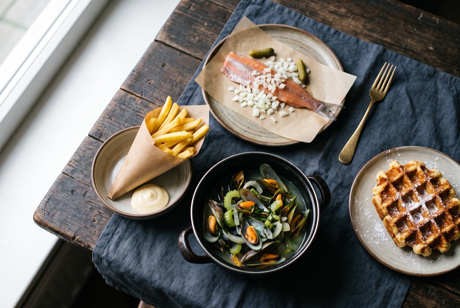 The Low Countries table: a wide dark wooden table seen from above, a pot of moules-frites with mussels and a small paper cone of fries, a raw Hollandse Nieuwe herring on a plate with chopped onion.