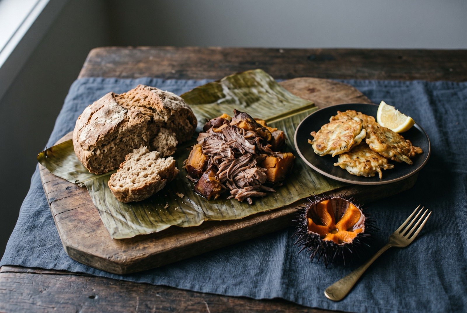 The Māori table: a wide dark wooden table seen from above, a torn loaf of rēwena bread beside a mound of shredded hāngī pork and kūmara on a banana leaf.