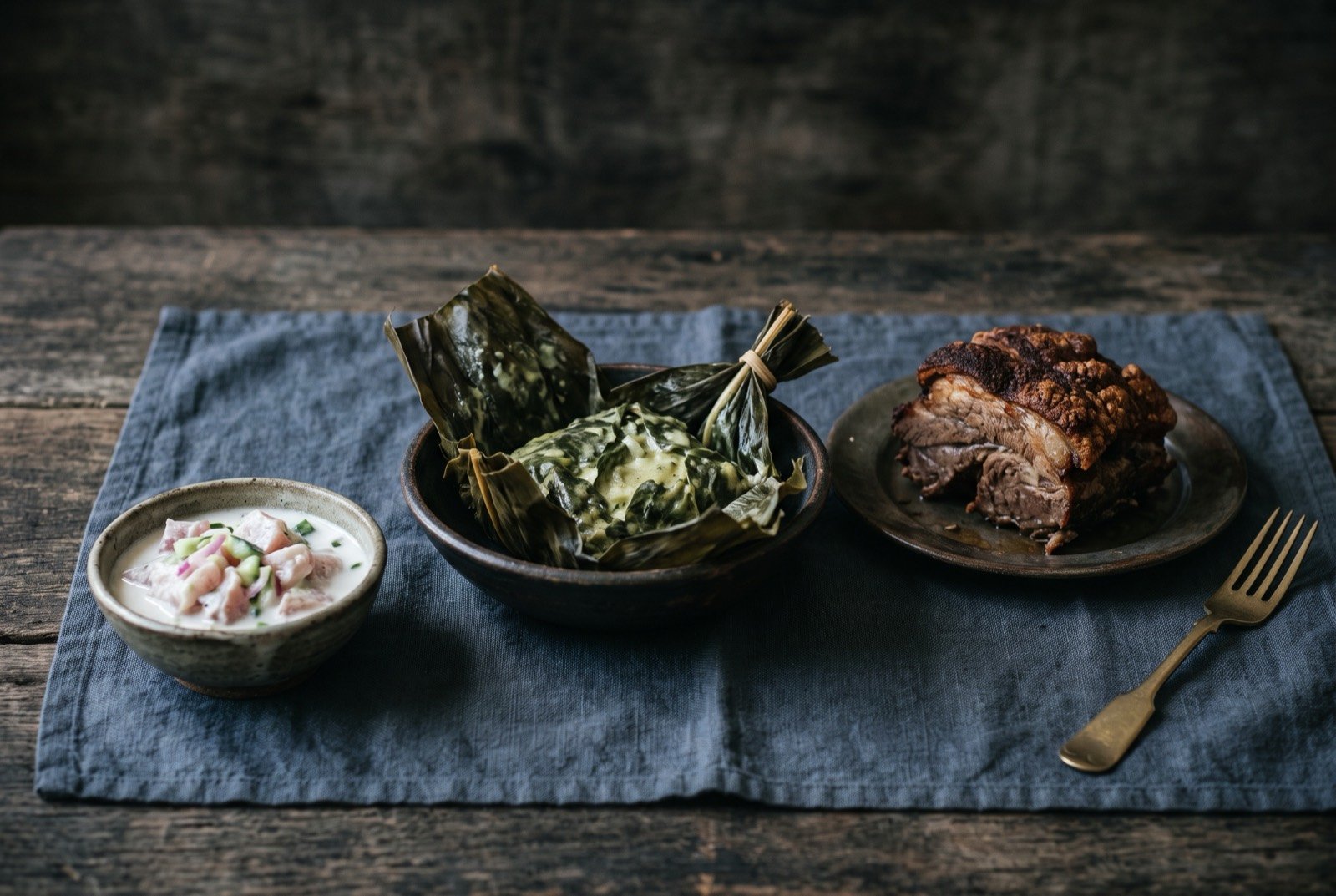 The Samoa, Tonga and Niue table: a wide dark wooden table seen from above, an opened parcel of palusami with coconut cream in taro leaf, a piece of umu-roasted pork.